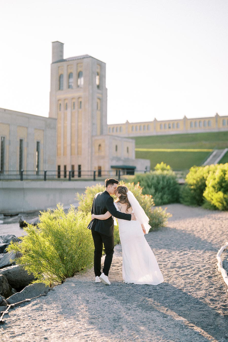 A bride and groom walking along a picturesque path by a historic building, surrounded by lush greenery on a sunny day.