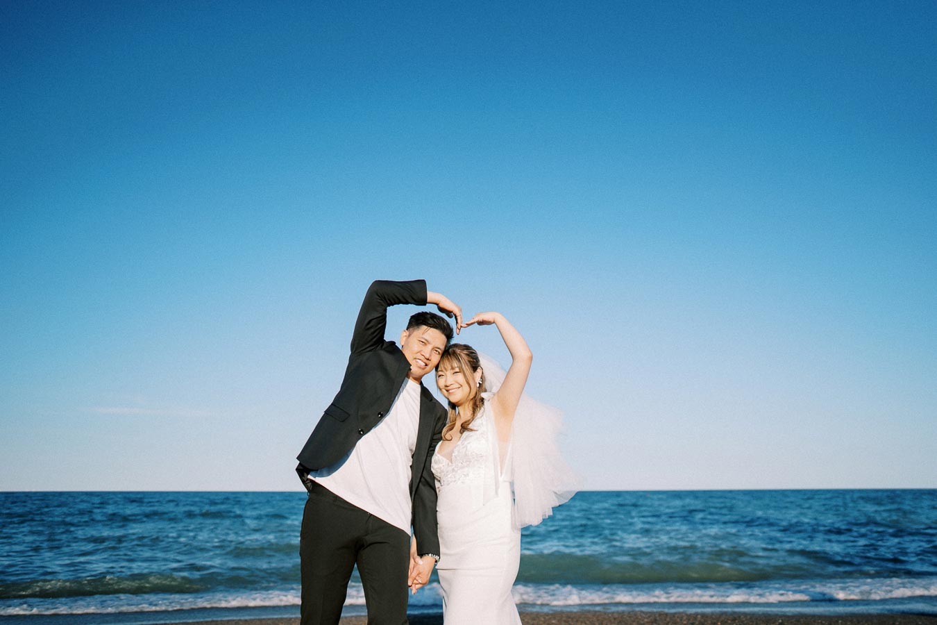 Happy couple posing in wedding attire by the ocean, creating a heart shape with their arms on a clear, sunny day.