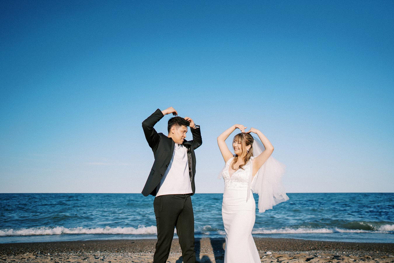 A bride and groom joyfully pose on a beach, forming heart shapes with their arms against a clear blue sky and ocean backdrop, capturing a playful wedding moment.