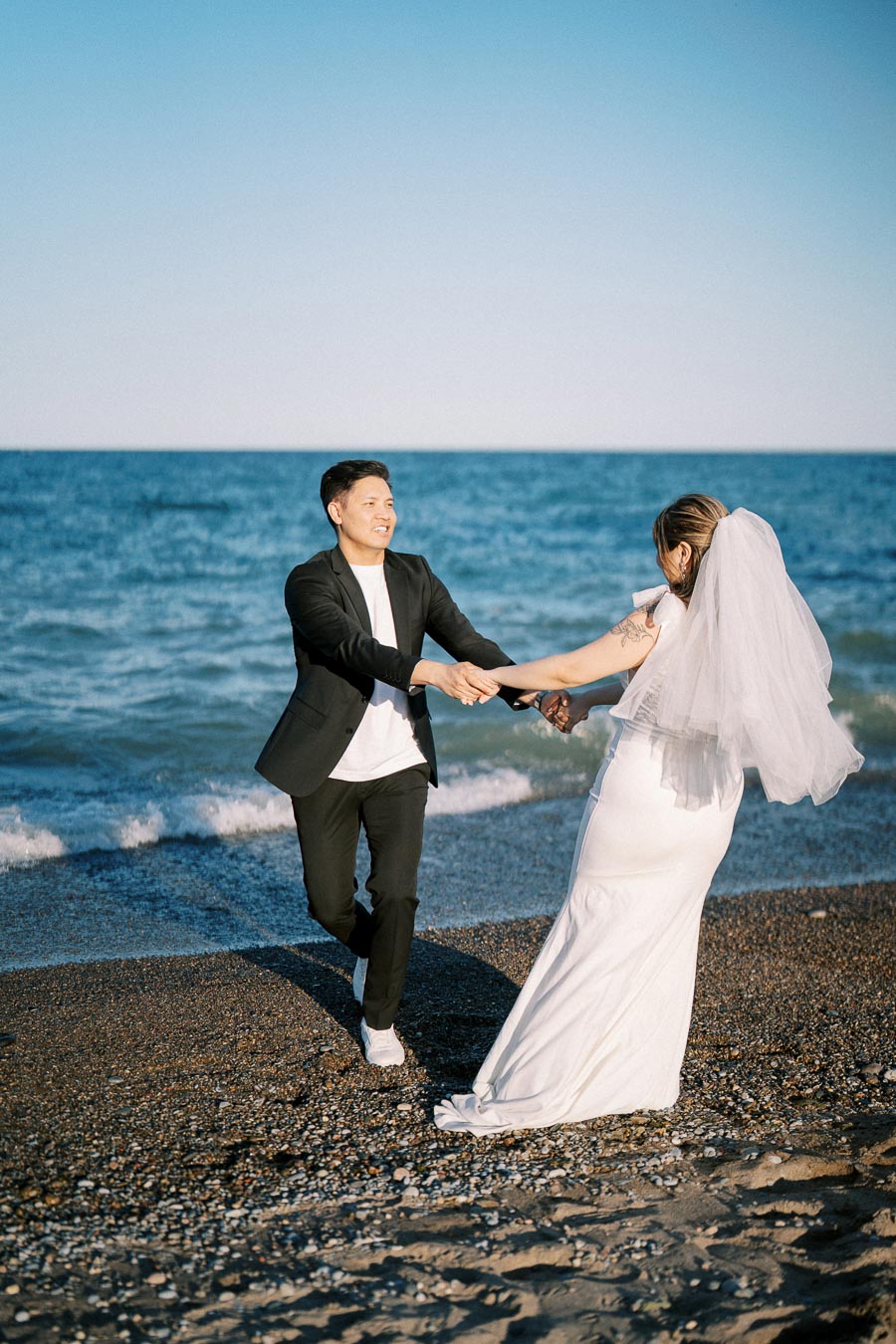 A bride and groom joyfully hold hands and dance on a sandy beach with the ocean in the background, under a clear blue sky.