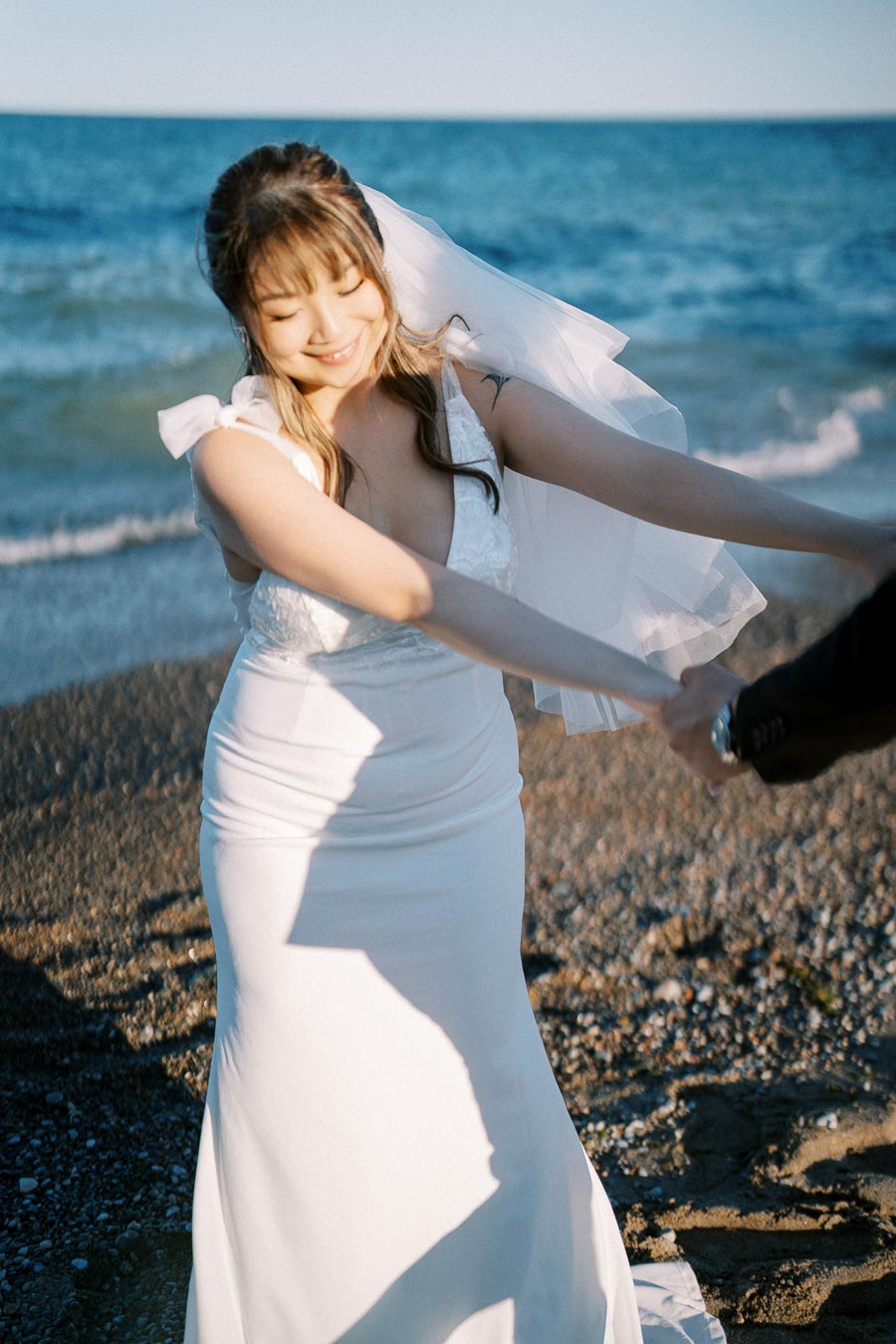 Bride in a white wedding dress smiling and holding hands on a sandy beach with ocean waves in the background, capturing a joyful seaside wedding moment.