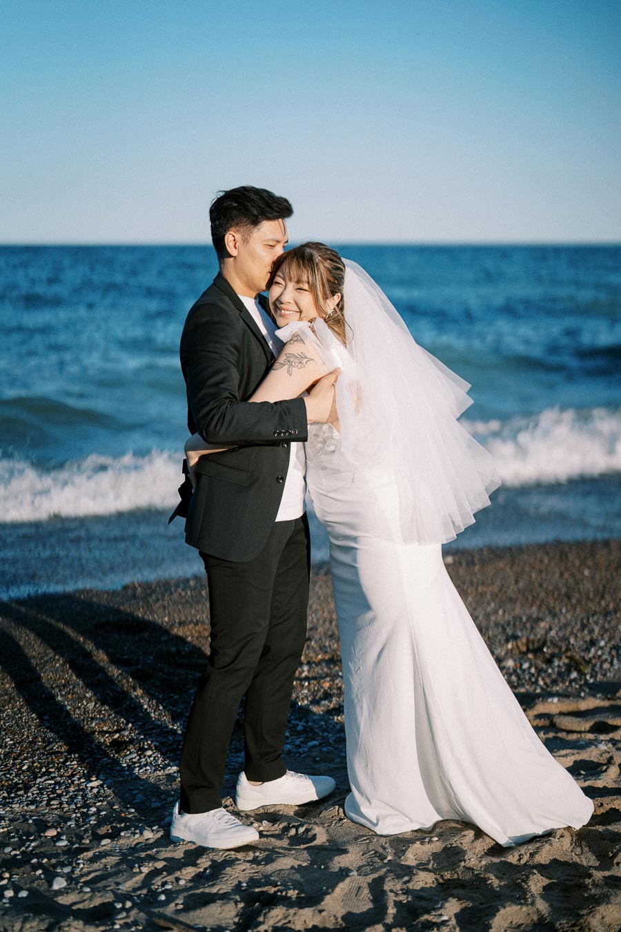 Happy couple in wedding attire embracing on a sandy beach with blue ocean waves in the background under clear skies.
