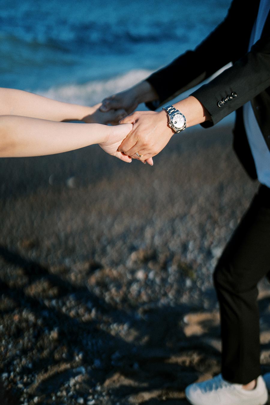 Two people holding hands on a sandy beach, one wearing a watch and the other a ring, with the ocean in the background.