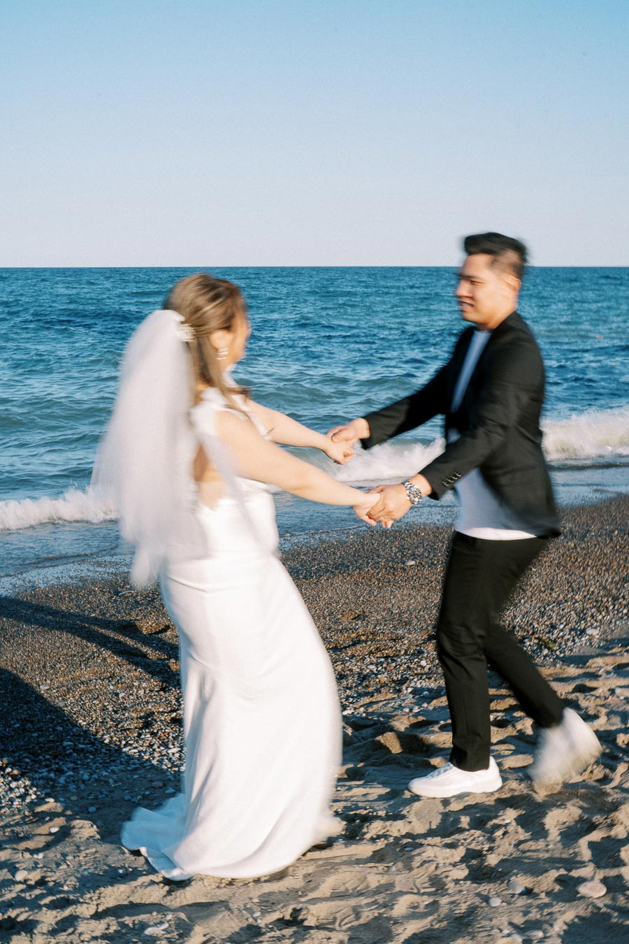 Happy couple in wedding attire dancing joyfully on a sandy beach by the ocean under a clear blue sky.