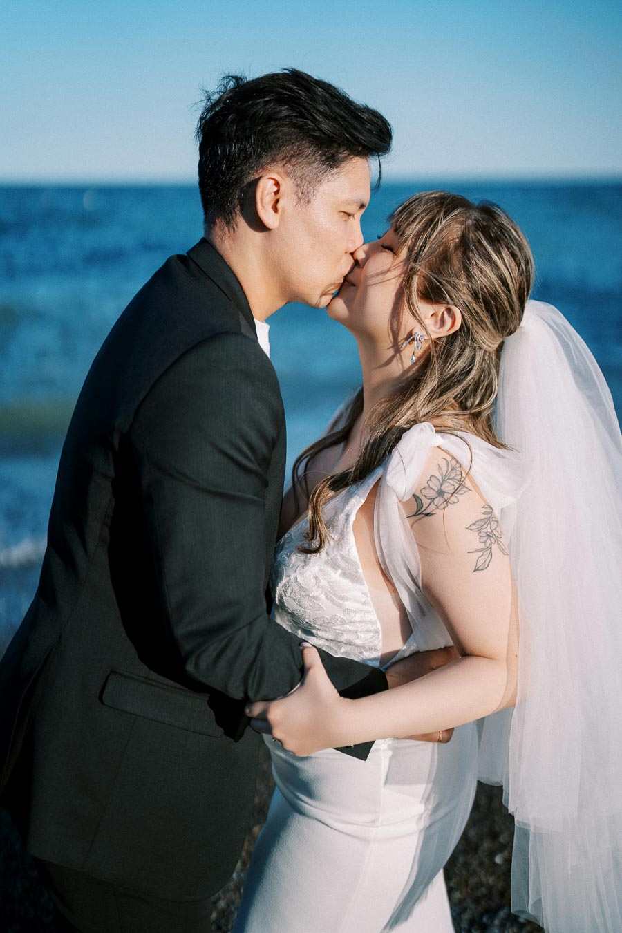 A newlywed couple sharing a romantic kiss on the beach, with the bride in a white dress and veil and the groom in a black suit, under clear blue skies.