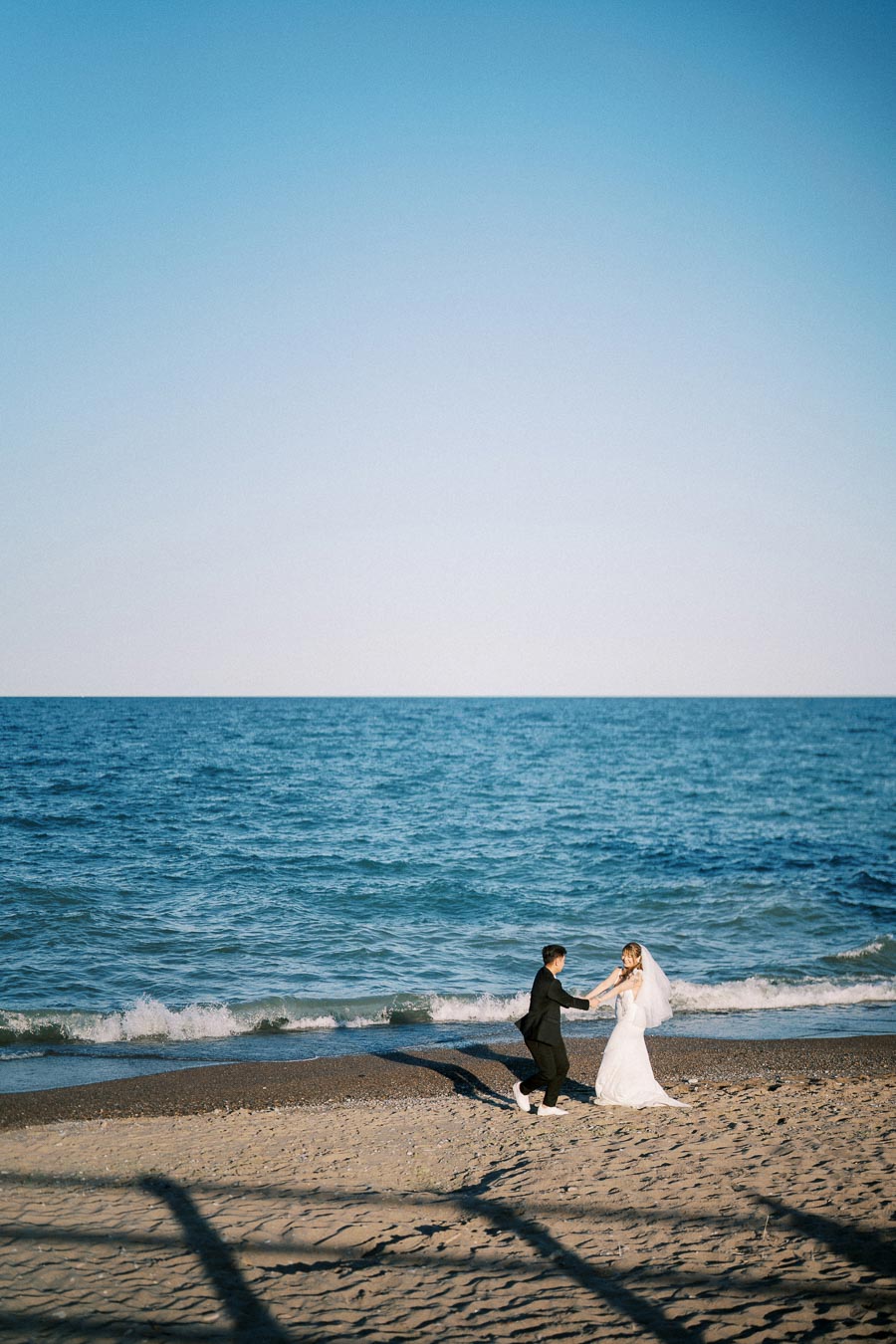 A couple in wedding attire joyfully holds hands on a sandy beach by the sea under a clear blue sky.