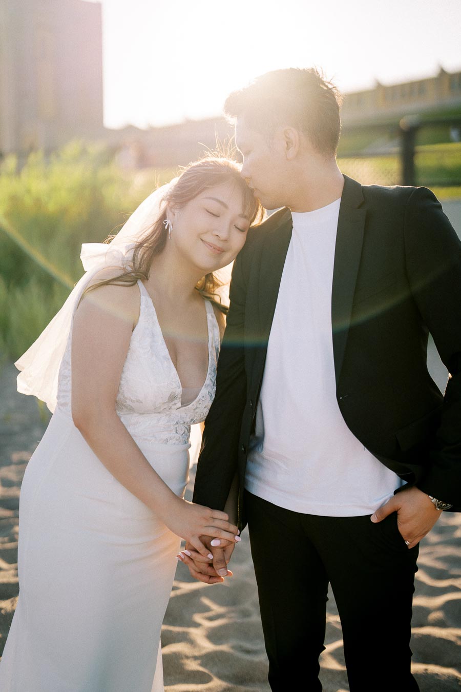 Romantic beach wedding photo of a couple holding hands, with the groom kissing the bride's forehead, bathed in warm sunset light.