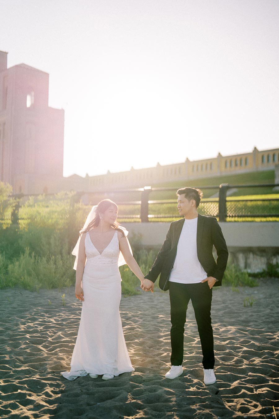 A couple in wedding attire holding hands and walking on a sandy path, with a historic building and greenery in the background during sunset.