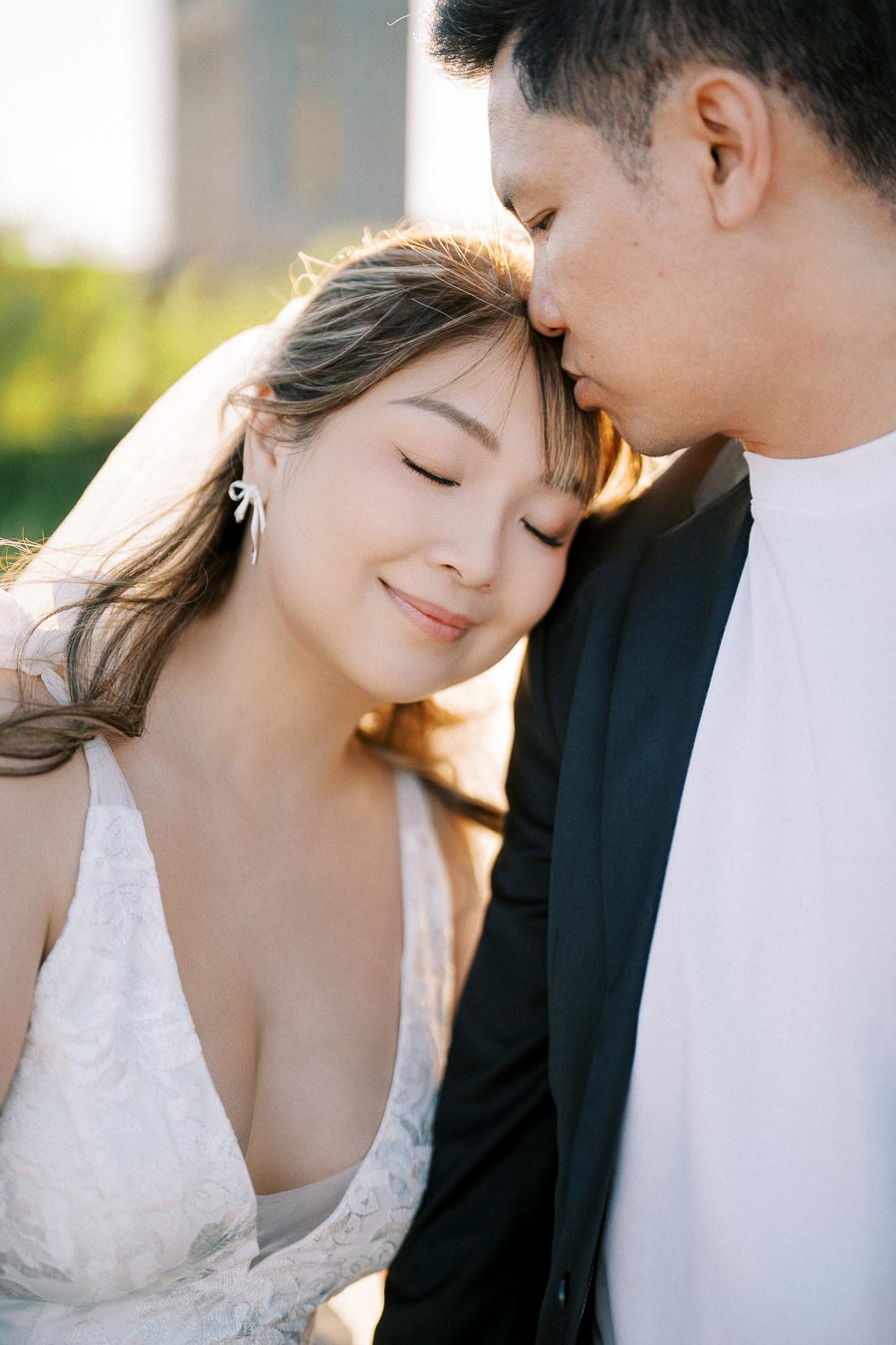 A couple embracing lovingly outdoors, with the woman smiling and the man gently kissing her forehead; they are dressed in elegant wedding attire, creating a romantic and serene atmosphere.