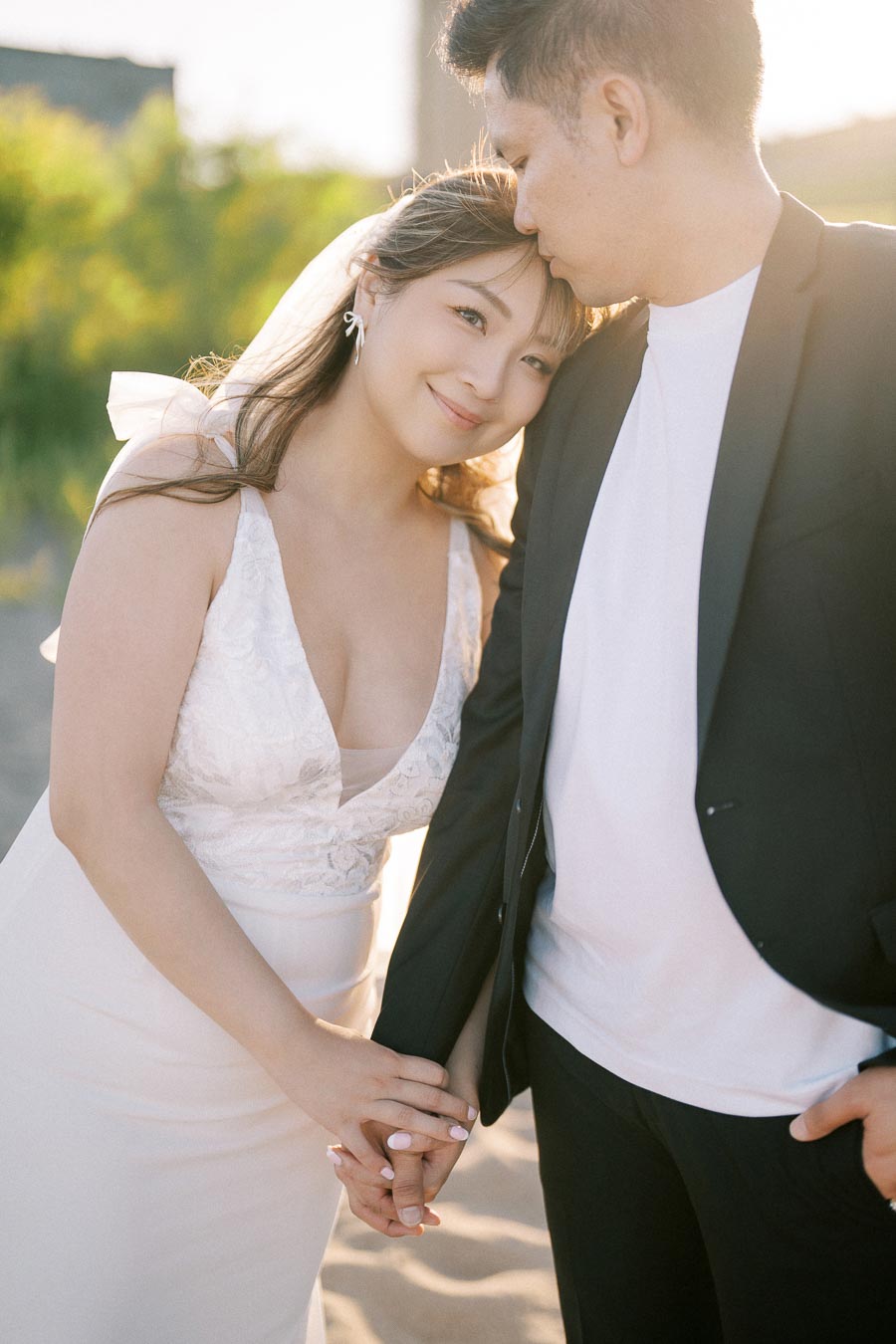 Couple celebrating wedding day, bride in elegant white dress and groom in black suit, holding hands and smiling lovingly at beach during sunset.