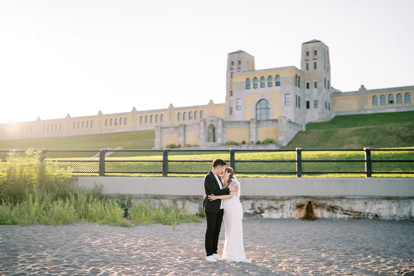 Bride and groom embracing on a sandy beach with a historic building in the background, bathed in warm sunset light.