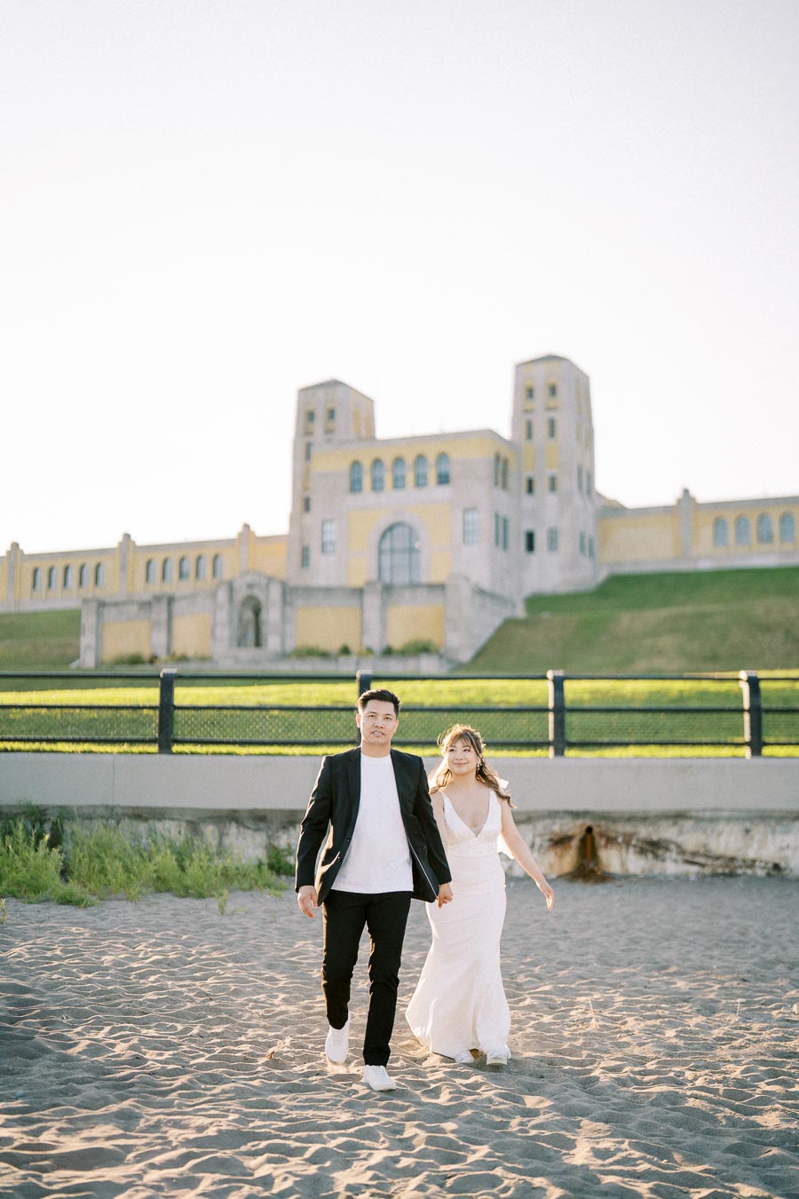 A couple walking hand in hand on a sandy beach with a large architectural building in the background, bathed in soft sunlight.