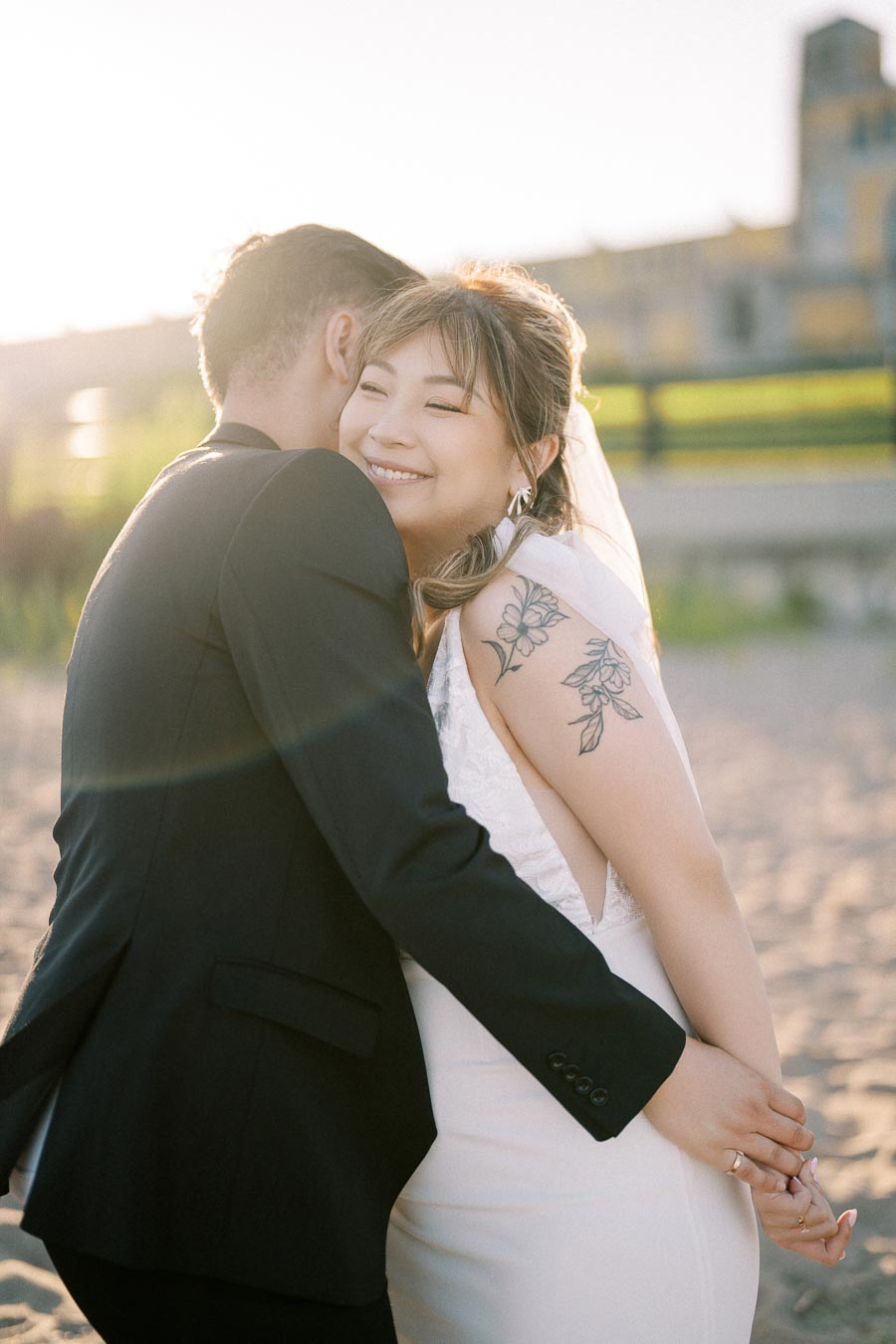 Smiling bride with floral tattoo embraces groom on sandy beach during sunny wedding day.