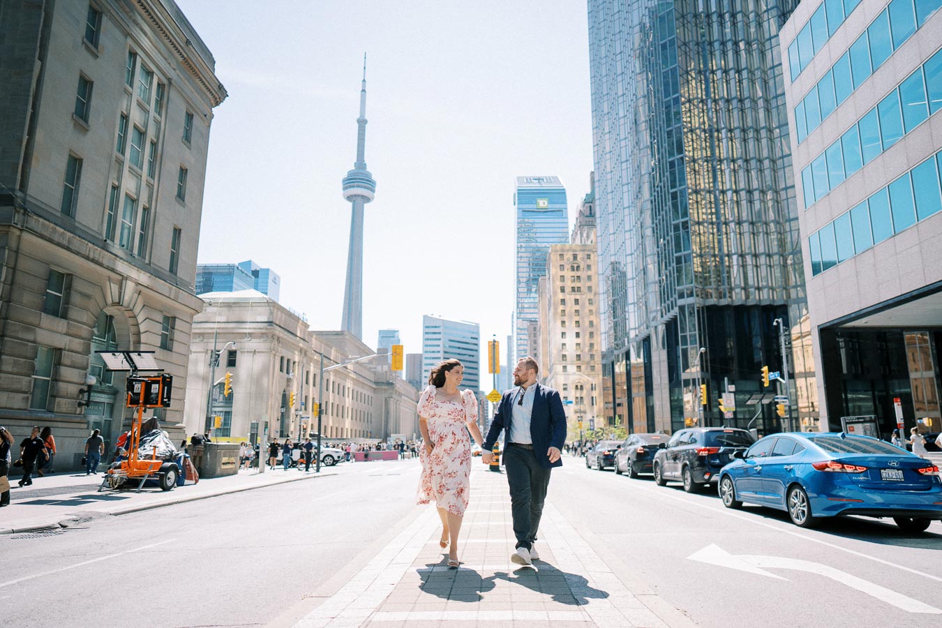 A couple walks hand in hand in downtown Toronto, with the CN Tower and modern skyscrapers in the background, under a clear blue sky.