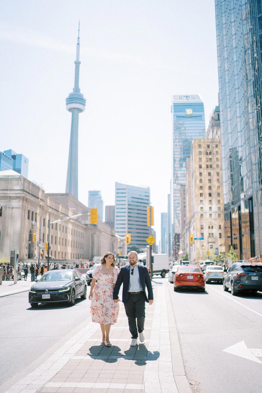 A couple walking hand in hand on a sunny day in a busy city street, with the CN Tower and tall buildings in Toronto, Canada in the background.