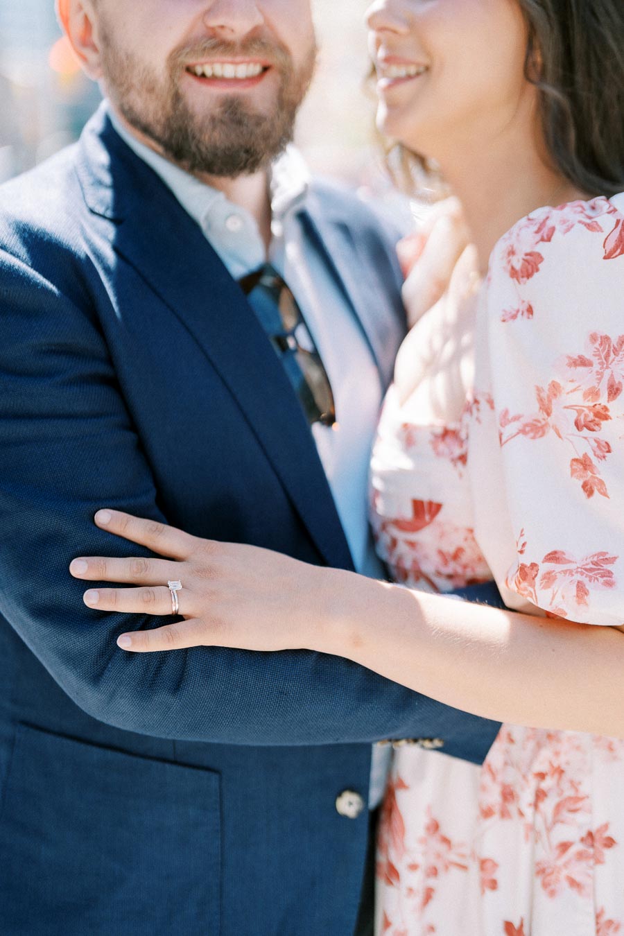 Couple embracing with woman showing engagement ring, man in blue suit and woman in floral dress.