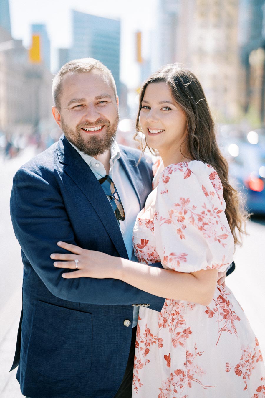 A smiling couple embracing in a sunny, urban setting, with modern buildings in the background. The man is wearing a blue blazer, and the woman is in a floral dress, highlighting a happy, romantic moment.