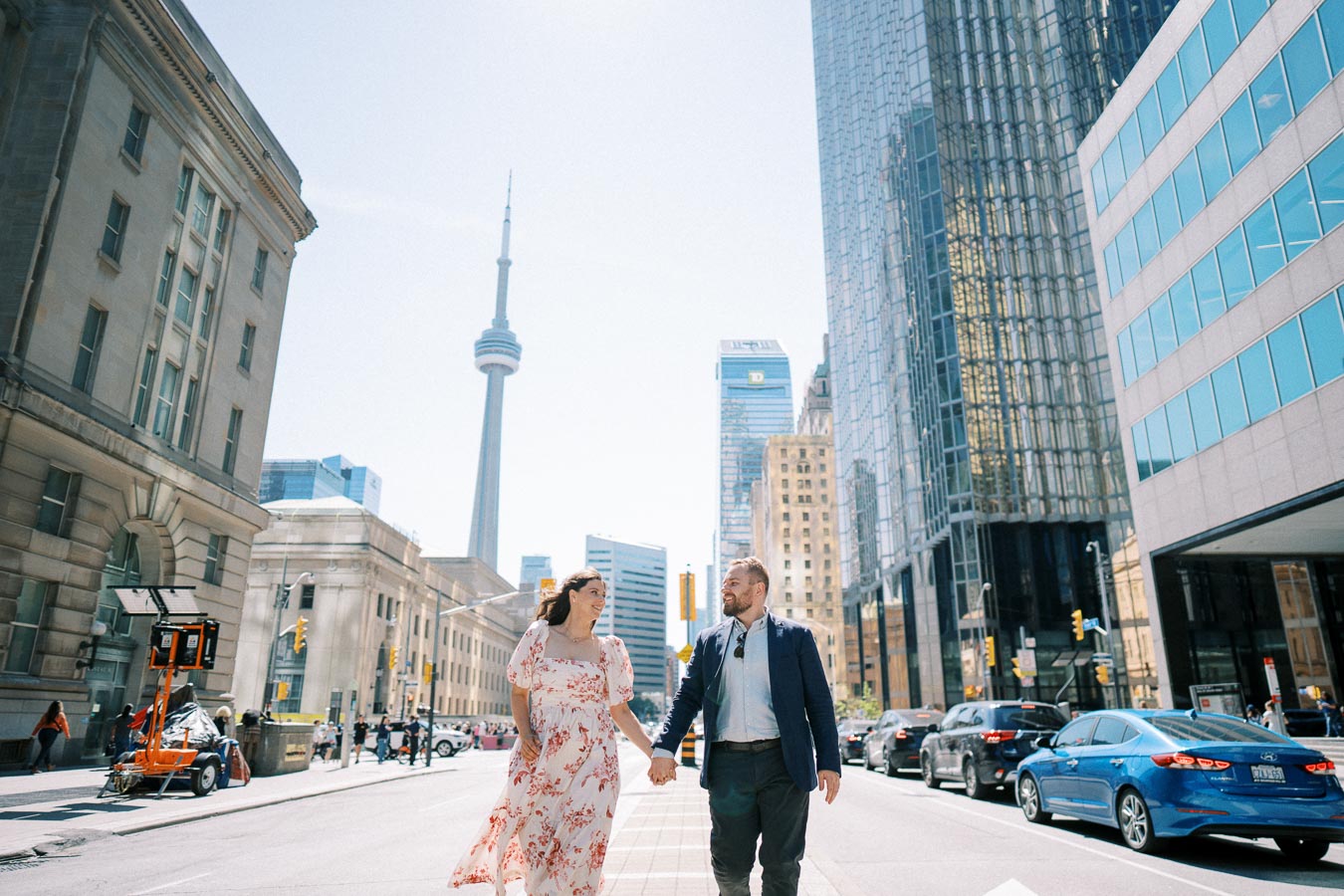 A couple holding hands and walking in downtown Toronto, with the CN Tower and modern skyscrapers in the background on a sunny day.