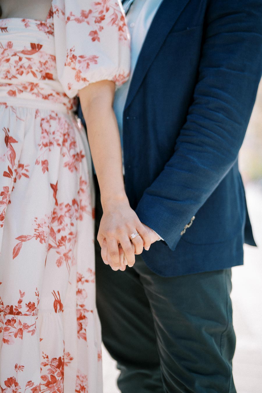 A close-up of a couple holding hands, with the woman wearing a floral dress and an engagement ring, and the man in a dark blue suit jacket.