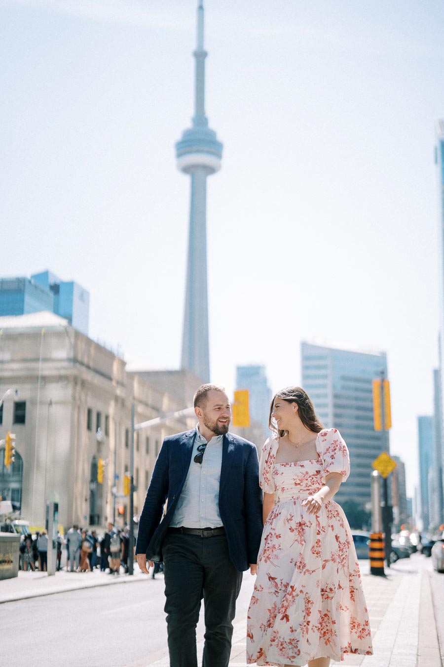 A couple smiling and walking together in a city with a tower in the background, capturing a joyful urban moment on a sunny day.