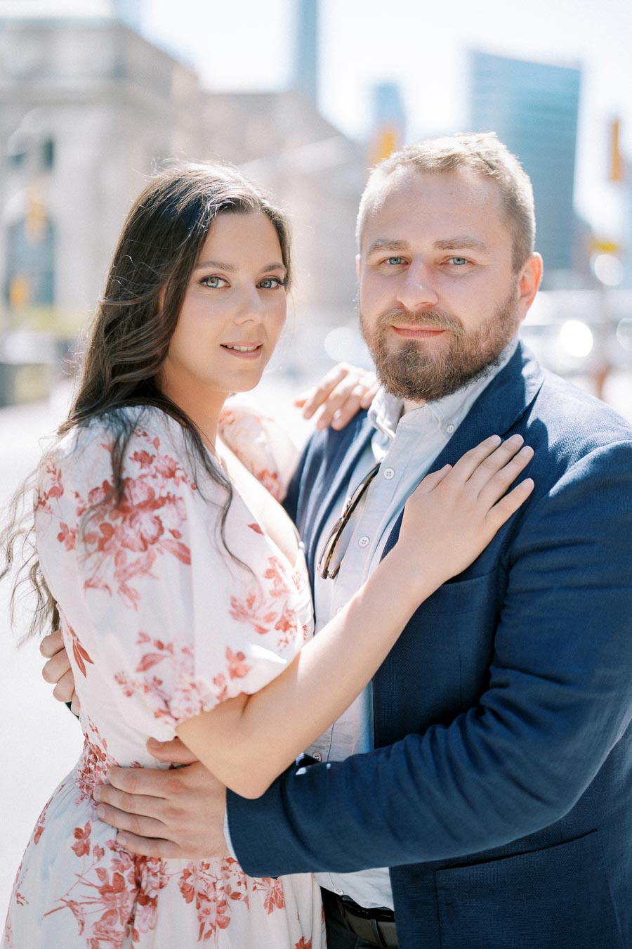 A couple embracing outdoors with a city skyline in the background, the woman wearing a floral dress and the man in a blue suit, both smiling at the camera.