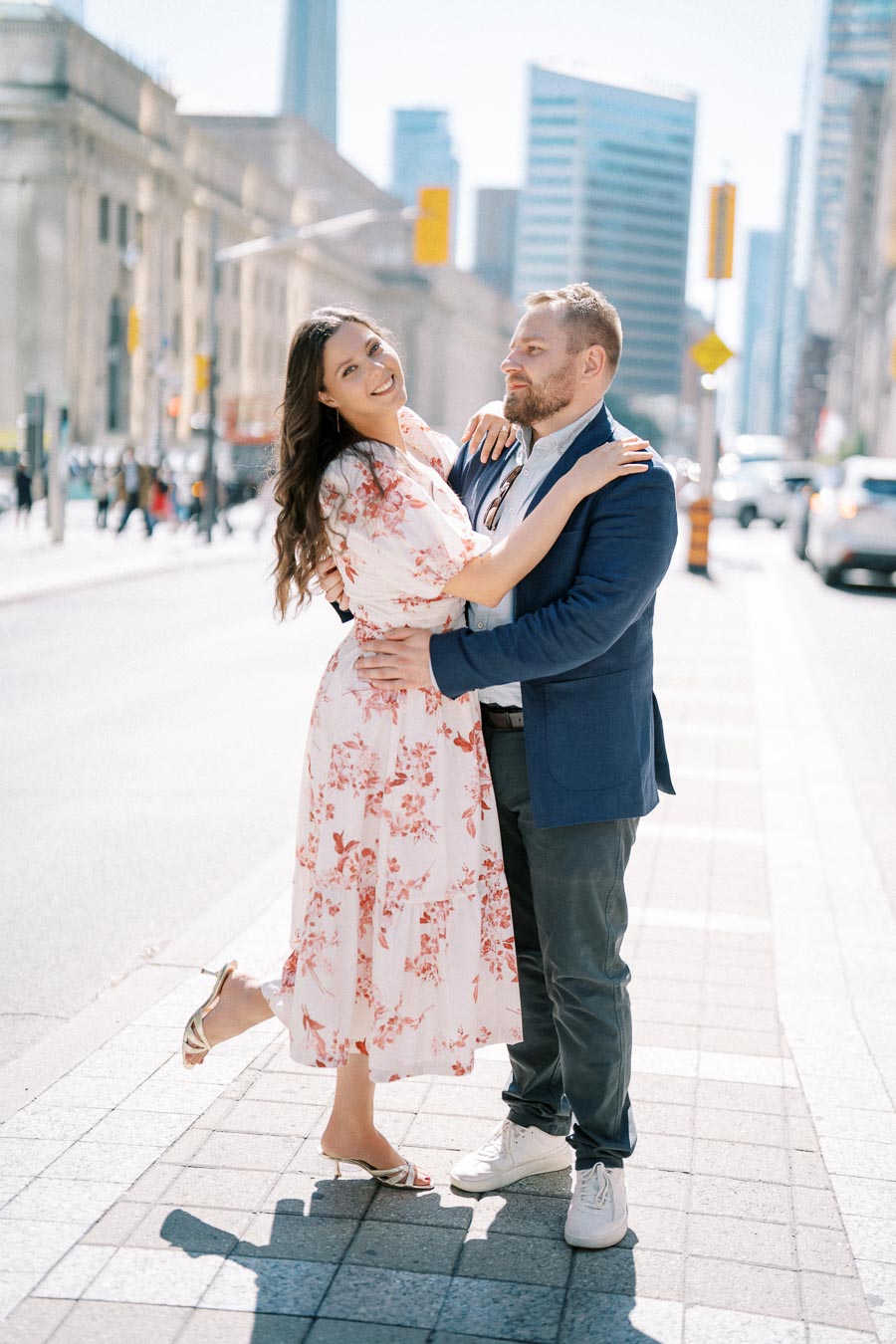 A happy couple embracing on a sunny city street, with modern buildings in the background. The woman is wearing a floral dress and heels, and the man is dressed in a blue jacket and sneakers.