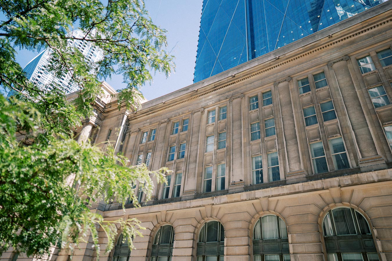 Historic stone building with tall windows and columns, framed by leafy green trees, with modern glass skyscraper in the background under a clear blue sky.