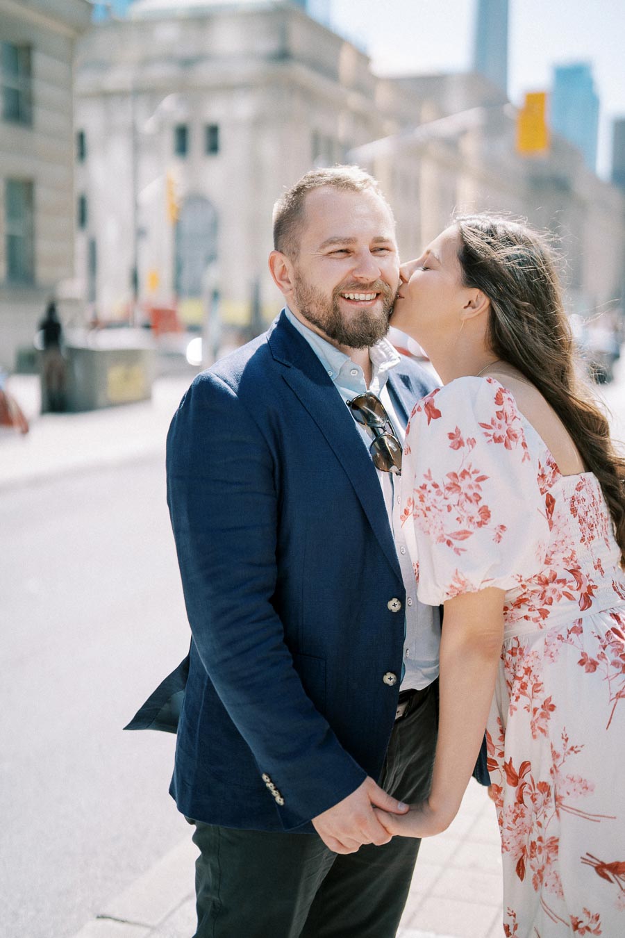 A couple standing on an urban street, the woman in a floral dress whispering to a smiling man in a blue jacket, holding hands.