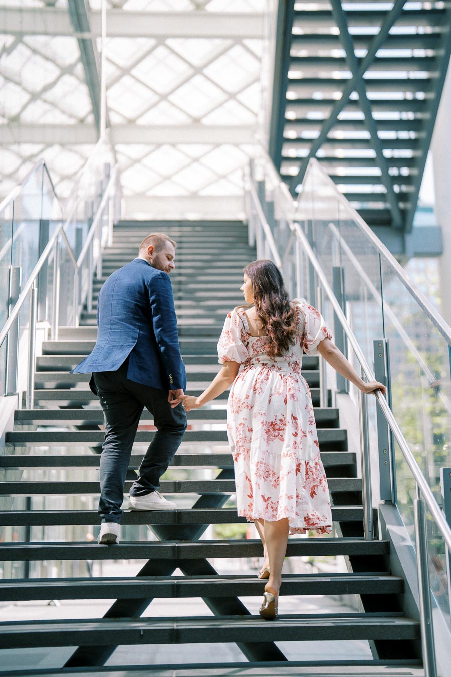 A couple holding hands and walking up a modern staircase, with glass railings and a geometric ceiling in the background, as sunlight filters through.