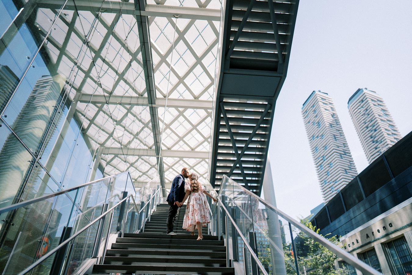 A couple holding hands and kissing on a modern glass staircase with skyscrapers in the background on a sunny day.