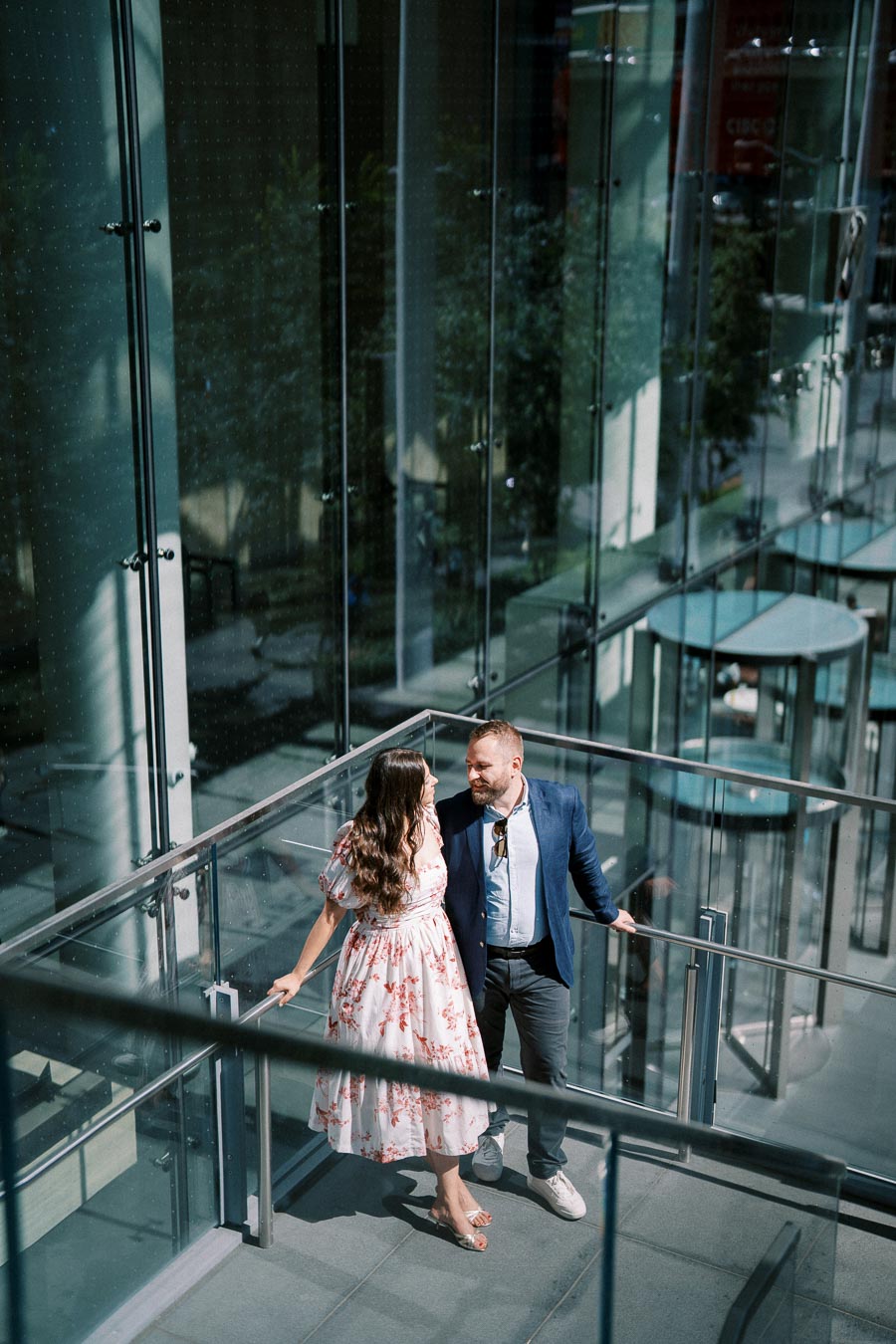 A couple standing on a glass staircase in a modern building, with the woman wearing a floral dress and the man in a blue suit jacket, looking at each other lovingly.
