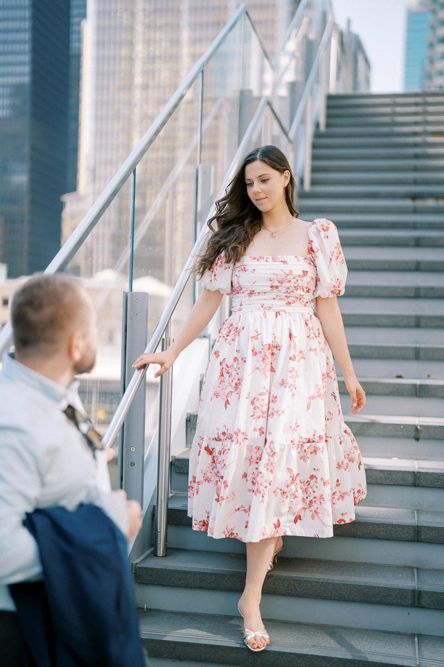 Young woman in a floral dress descends outdoor stairs on a sunny day, with modern city buildings in the background.
