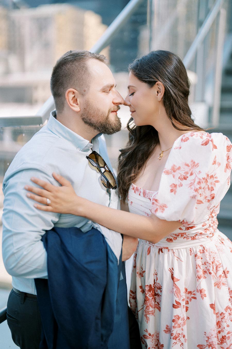 A couple sharing a tender moment on a staircase, with the woman in a floral dress and the man holding a jacket, embracing closely in an urban setting.