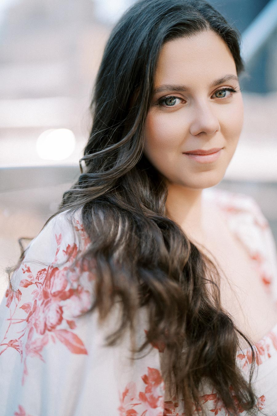 Close-up portrait of a woman with long dark hair and a floral patterned outfit, smiling softly in an outdoor setting.