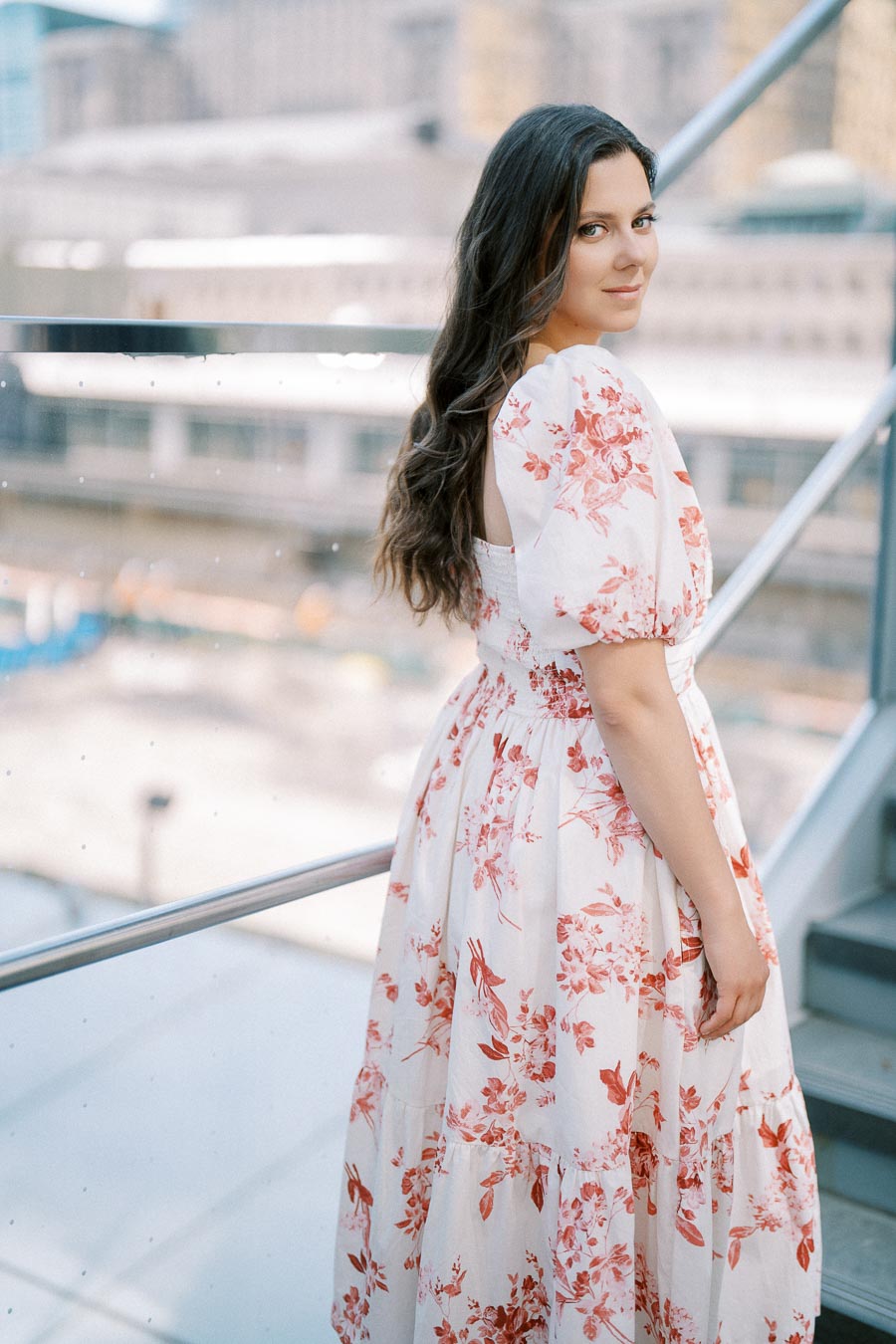 A woman in a floral dress stands by a glass railing with an urban backdrop.