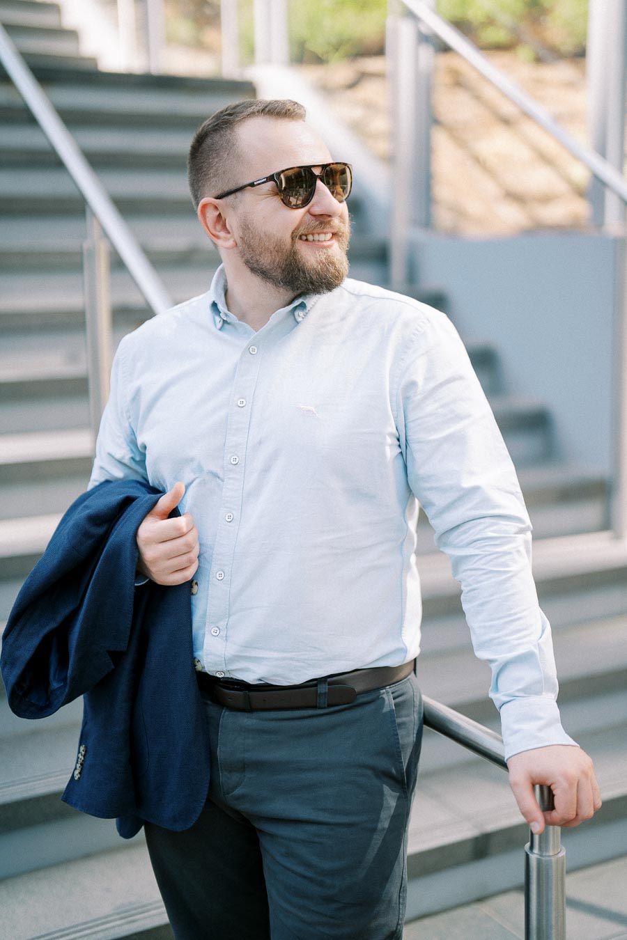 Man wearing sunglasses holding a blue jacket while standing on a modern staircase in a bright urban setting.