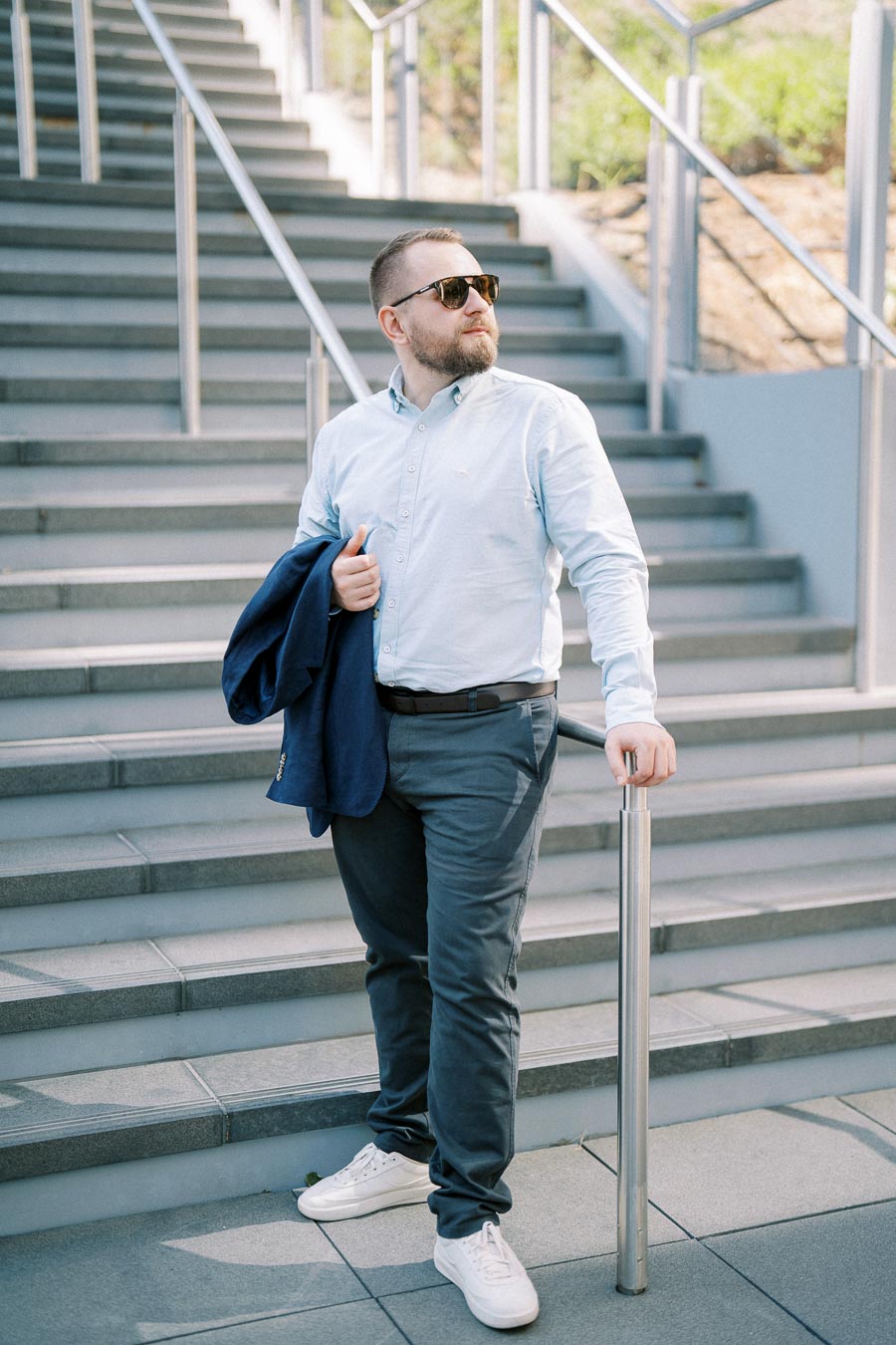 Man with sunglasses holding a jacket, standing on modern outdoor staircase.