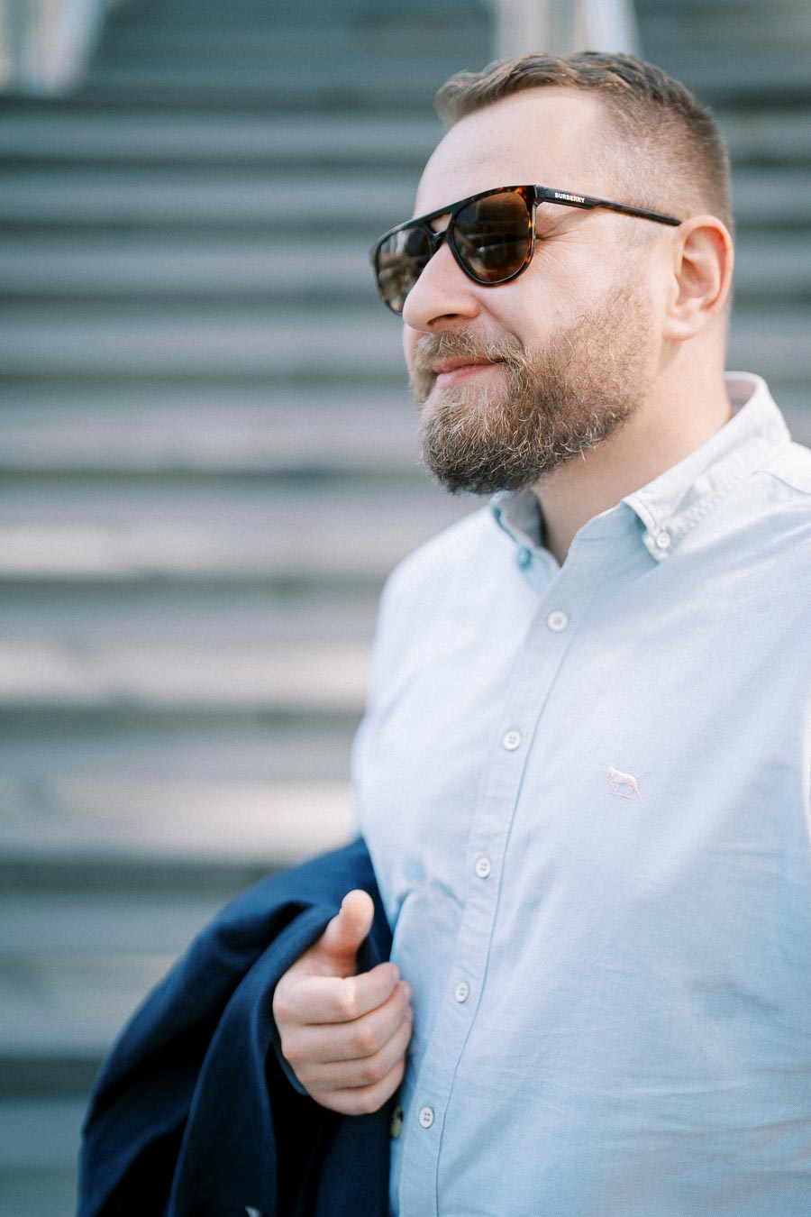 A stylish man in sunglasses and a light blue shirt holds a dark jacket while confidently standing on stairs outdoors.