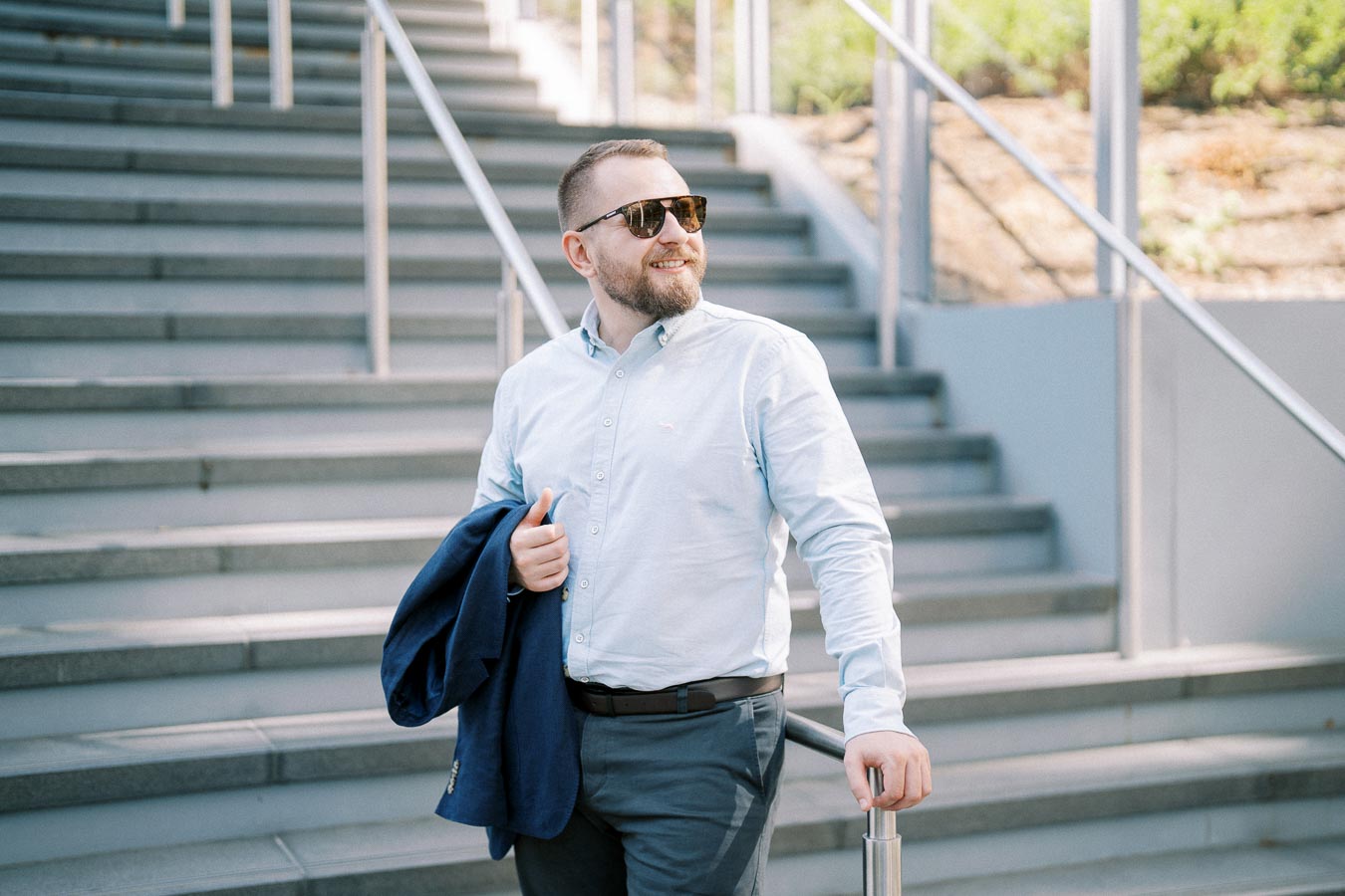 Man in sunglasses holding a jacket, confidently walking down stairs outdoors.