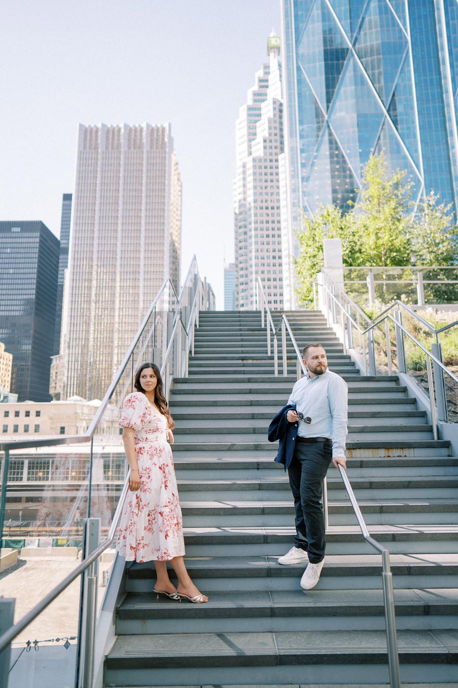 A man and woman standing on a staircase in an urban setting, surrounded by modern skyscrapers and greenery, on a clear day.