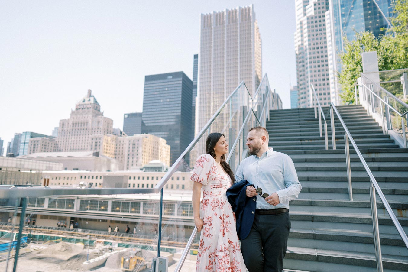 A couple walking down stairs with a modern city skyline in the background. The woman is wearing a floral dress and the man is in a light blue shirt, holding a jacket. Urban setting with skyscrapers and construction site below.