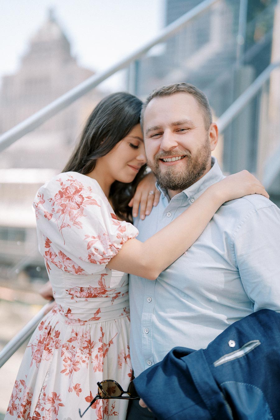 A happy couple embracing outdoors, with the woman in a floral dress and the man in a light blue shirt, both smiling contentedly in a city setting.