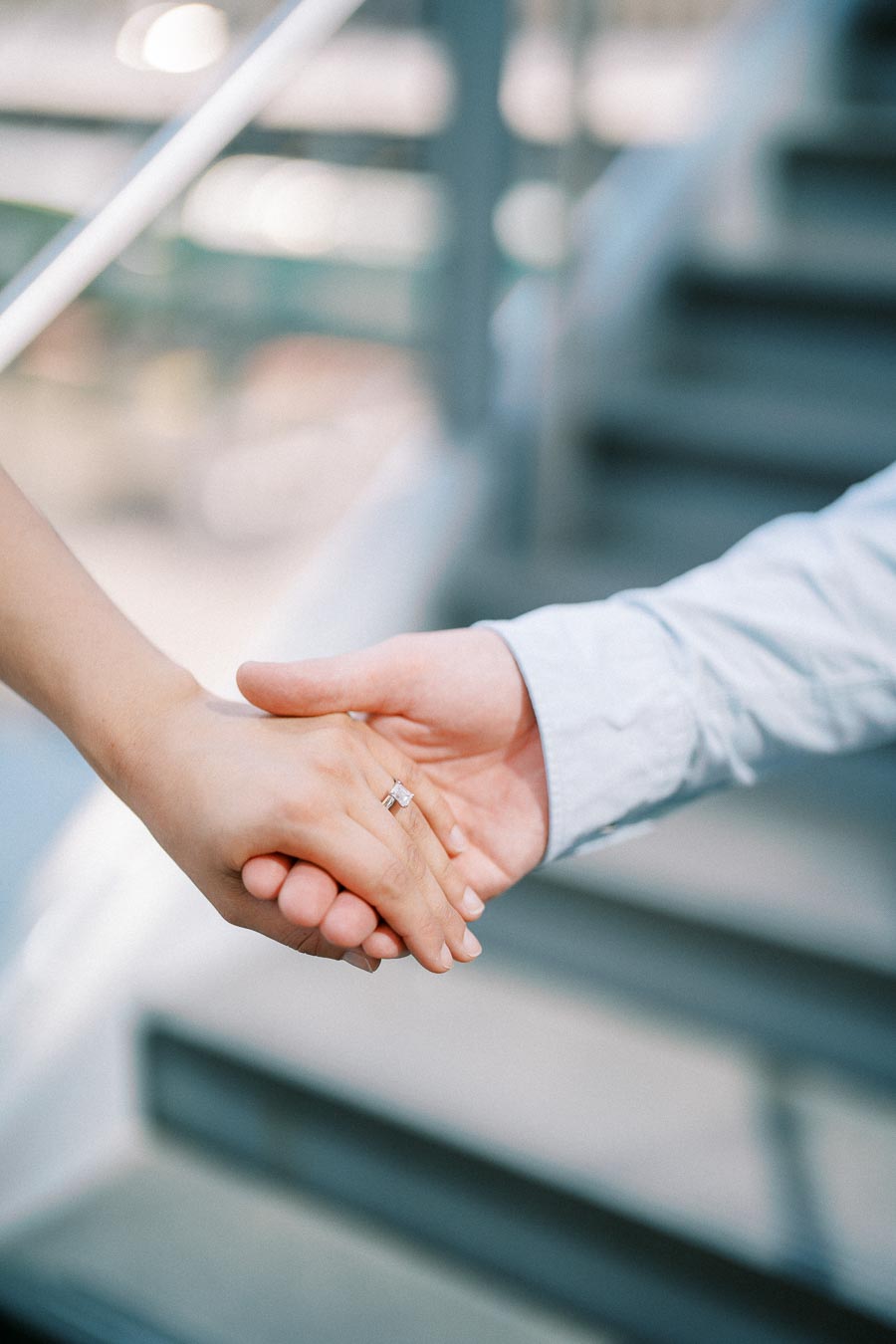 Two people gently holding hands, one wearing an engagement ring, symbolizing love and commitment. Blurred staircase in the background.