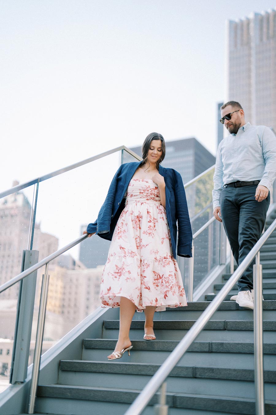 A woman in a floral dress and blue jacket descends outdoor stairs, accompanied by a man in a light blue shirt and sunglasses, in an urban setting with skyscrapers in the background.