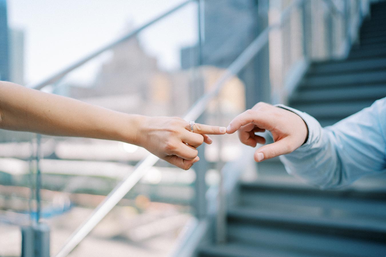 Two hands reaching towards each other, one with a ring, set against a blurred urban background with stairs, symbolizing connection and engagement.