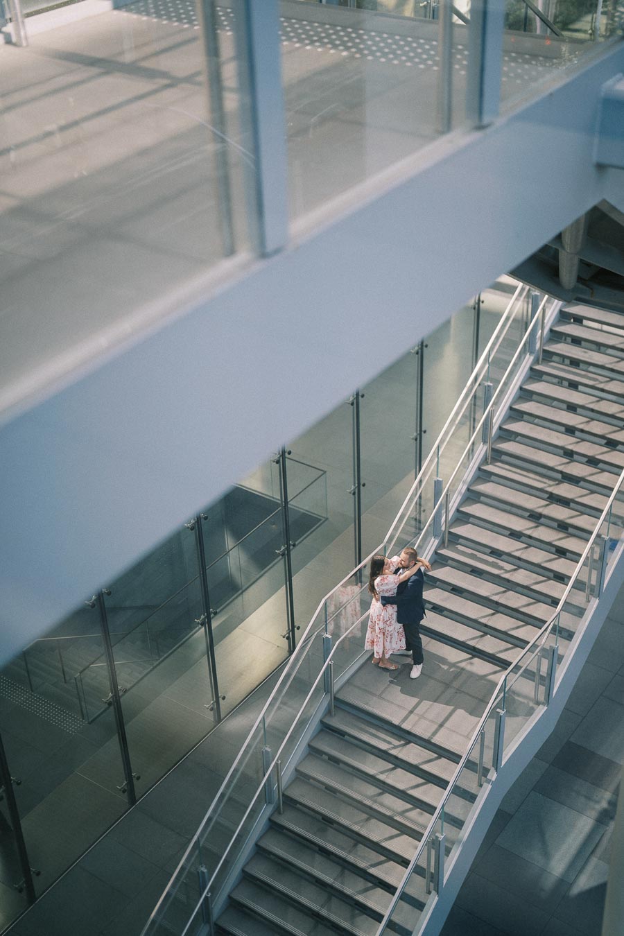 A couple embracing on a modern glass and steel staircase, captured from above, in a contemporary urban setting.