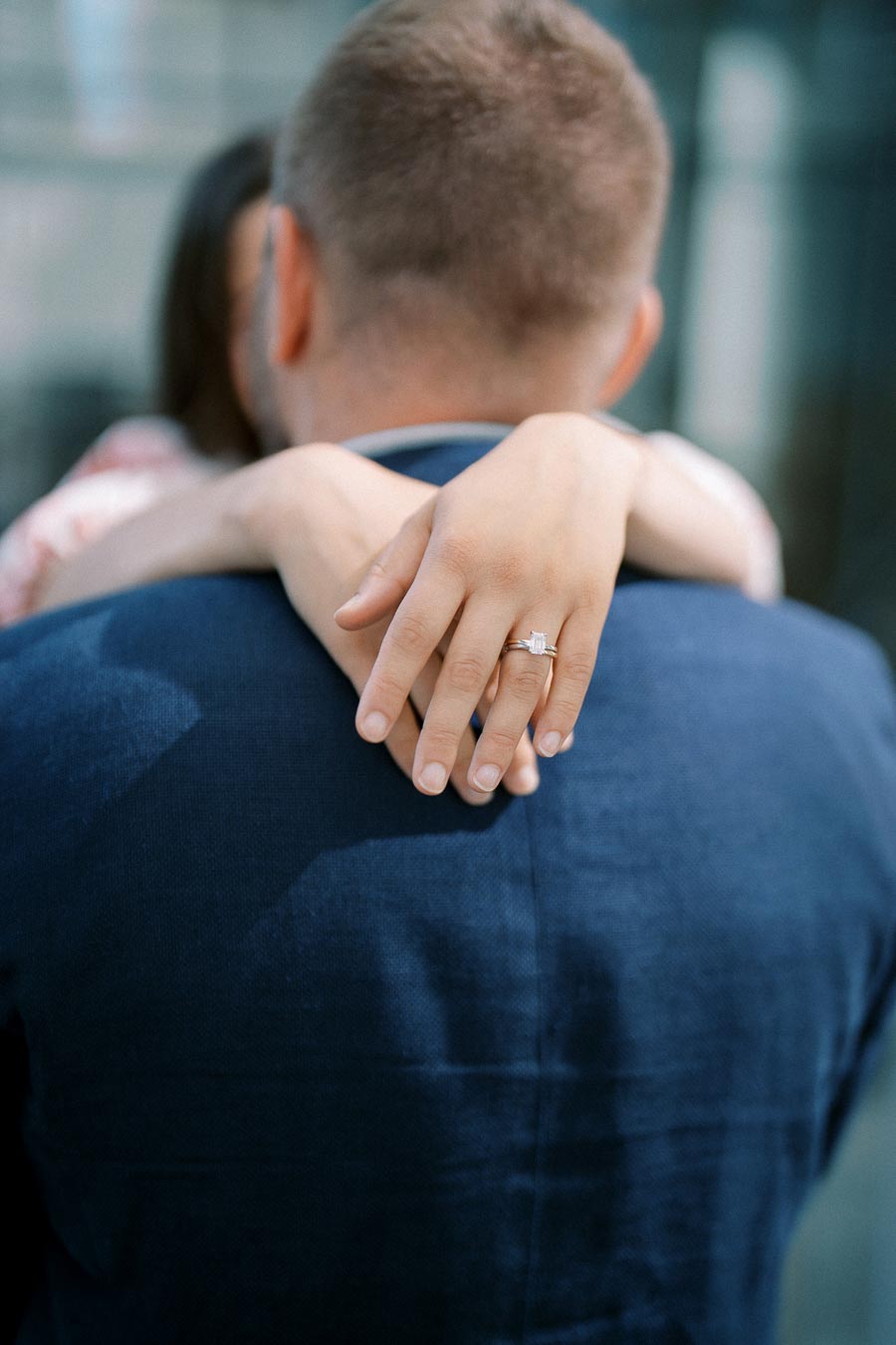 A woman's hand with an engagement ring resting on a man's shoulder, both wearing elegant clothing.