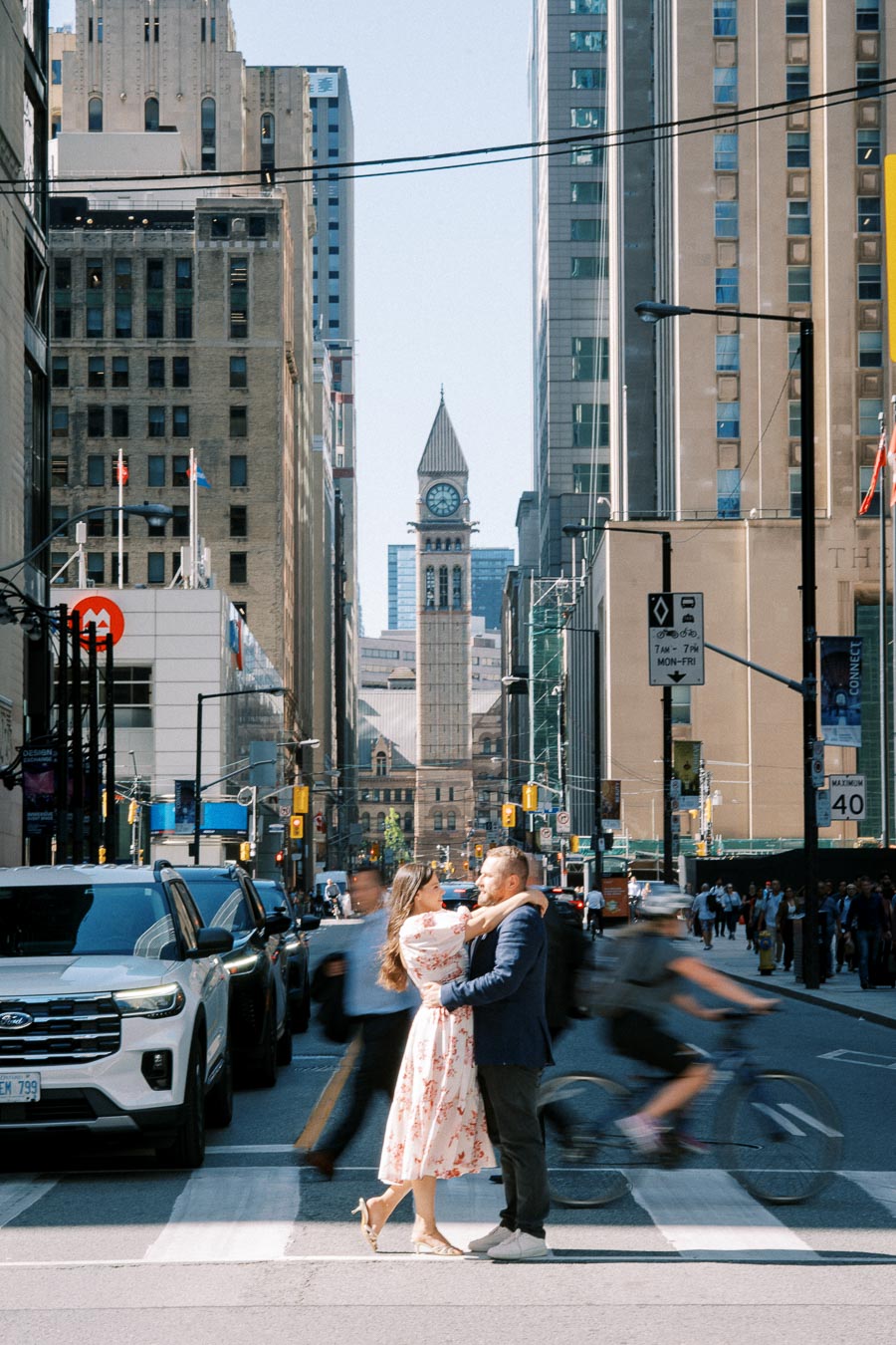 Couple embraces on busy Toronto street with iconic clock tower and tall buildings in the background. Urban romance and city life captured in vibrant downtown scene.