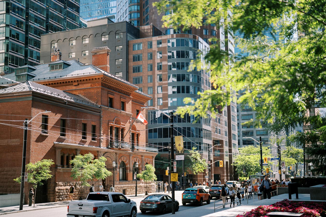 Urban street scene in Toronto with a historic brick building displaying a Canadian flag. Modern skyscrapers in the background, cars and pedestrians on the street, and greenery from trees under sunlight.