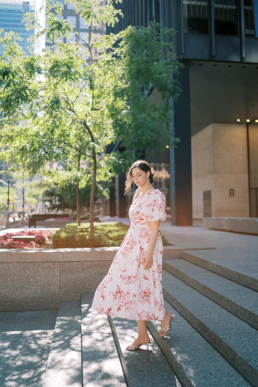 A woman in a floral dress stands gracefully on city steps surrounded by greenery, with sunlight filtering through the trees.